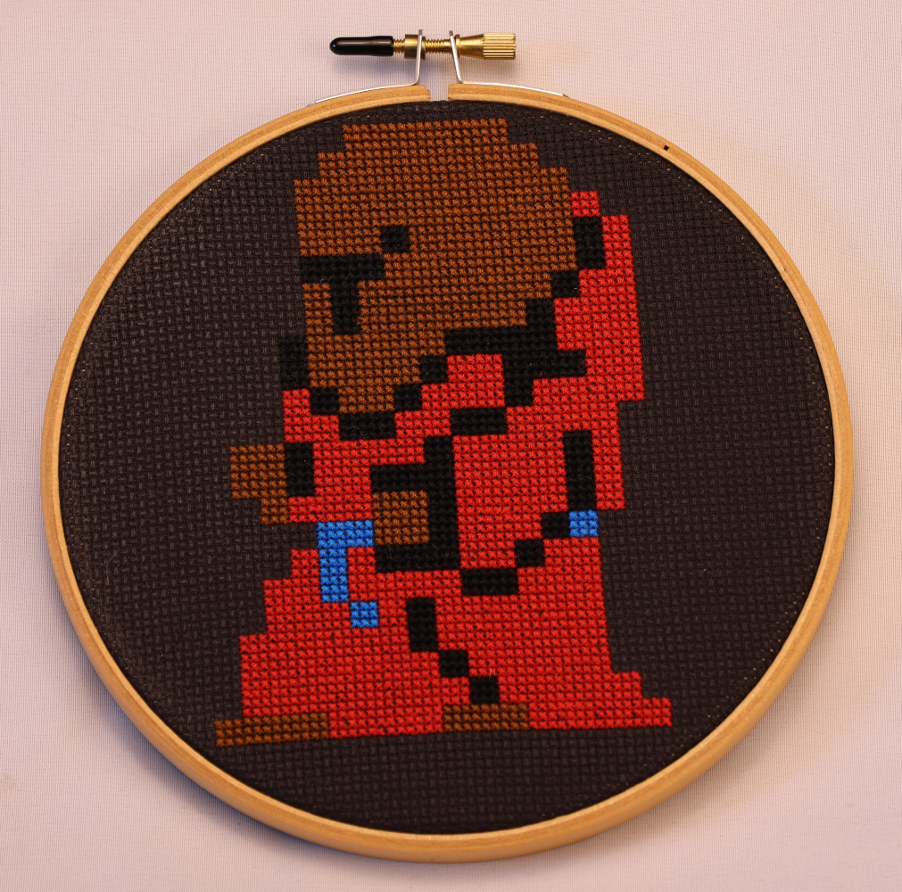 Photograph of a cross stitch on black aida. A pixel-rendered psion in the FF1 style with bald head, bare feet, red robes, a blue sash, and brown skin.